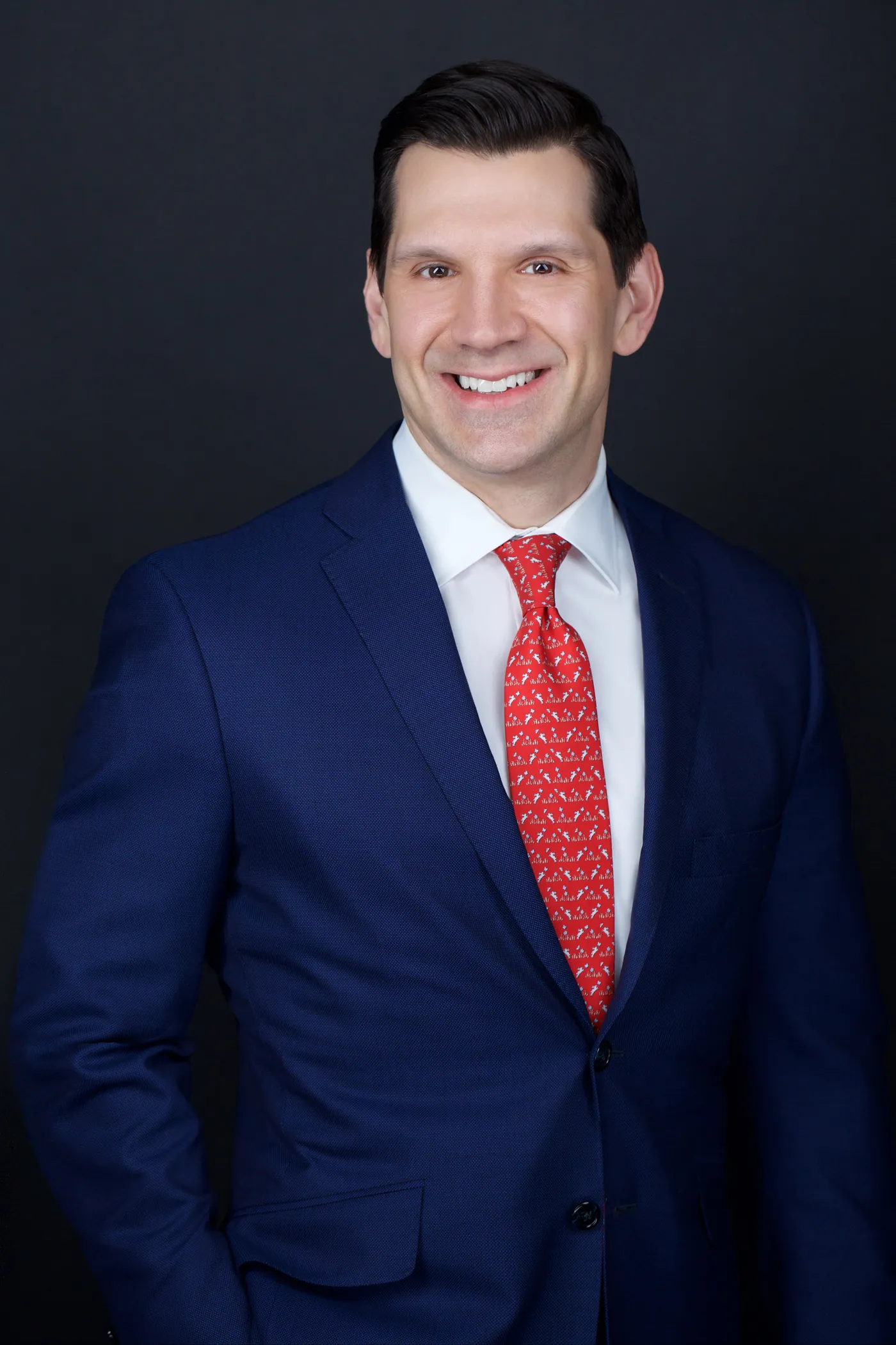 Corporate team headshot of a man in a navy suit and red tie, dark studio backdrop